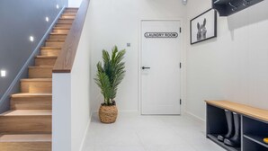 Un pasillo moderno con una escalera de madera, una planta en maceta, una puerta con la etiqueta 'Cuarto de Lavandería' y un banco con espacio de almacenamiento.