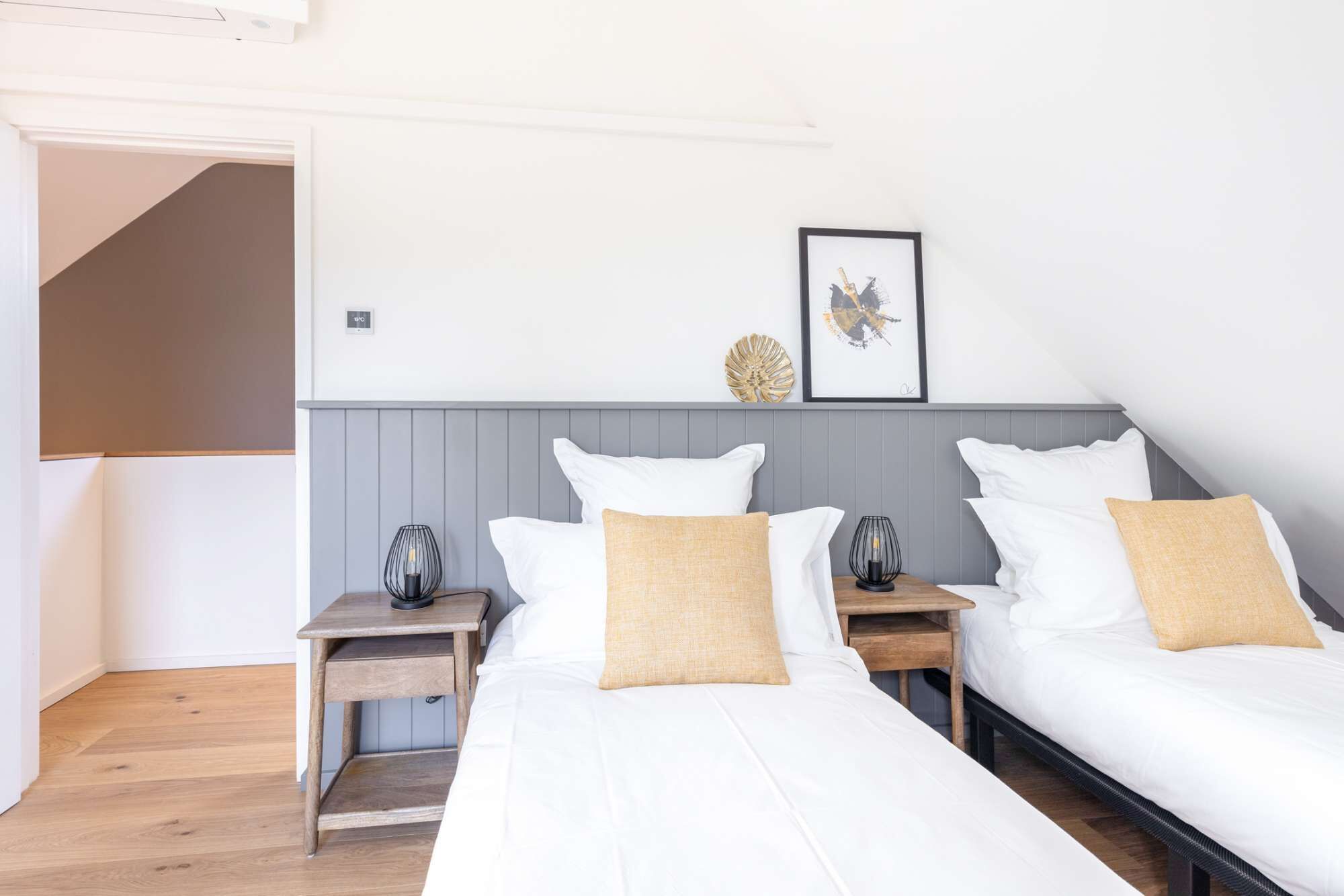 A hotel room with two single beds, a grey headboard, a small table with a lamp, and a framed picture on the wall.