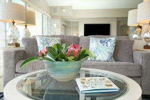 Uma sala de estar com uma mesa de centro de vidro, um livro intitulado "The Beachcomber's Companion" e um arranjo floral.