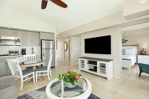 Uma cozinha moderna com eletrodomésticos de aço inoxidável, uma sala de jantar com cadeiras brancas, uma TV de tela plana na parede e uma sala de estar aconchegante com uma mesa de centro e um vaso de flores.