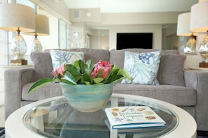 A living room with a glass coffee table, a book titled "The Beachcomber's Companion," and a floral arrangement.