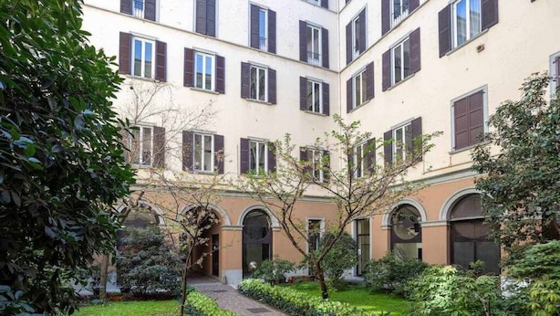 Une cour avec un chemin pavé, entourée de verdure et bordée d’un bâtiment doté de nombreuses fenêtres et de balcons.