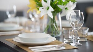 Uma mesa de jantar posta com pratos brancos, copos transparentes e um vaso com flores brancas.
