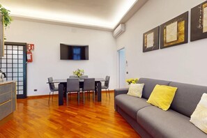 A modern living room with a grey sofa, wooden flooring, a dining table with chairs, and a mounted flat-screen TV.