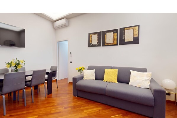 A modern living room with a grey sofa, a dining table with chairs, and artwork on the wall.