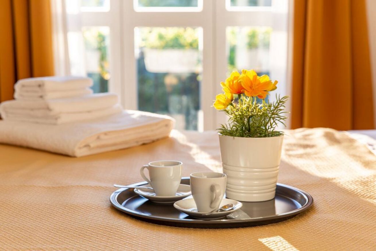 Un plateau avec une tasse blanche, une tasse plus petite et une plante en pot aux fleurs jaunes. En arrière-plan, on aperçoit un lit avec une pile de serviettes blanches soigneusement pliées.