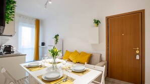 Un comedor con una mesa blanca dispuesta para cuatro personas, individuales amarillos y un florero con flores. Se observa una puerta de madera y un sofá beige con cojines amarillos al fondo.