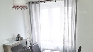 Een eethoek met een tafel met glazen blad, donkere stoelen, een klein kastje en een raam met dunne gordijnen.