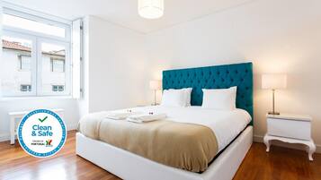 Habitación de hotel limpia y segura con cama blanca, cabecera color aguamarina y ventana con vistas a edificios.
