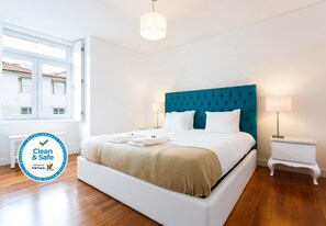 Une chambre d’hôtel propre et sûre, avec un lit blanc, une tête de lit turquoise et une fenêtre donnant sur des immeubles.