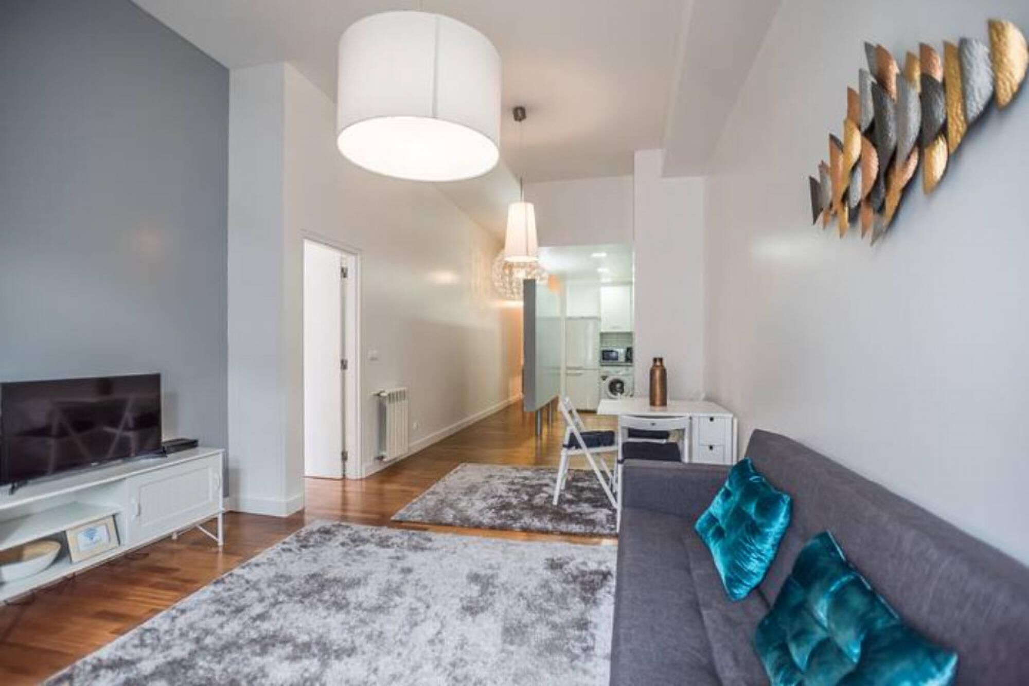 Una sala moderna con un sofá gris, un televisor y una mesita de centro. Hay un pasillo que conduce a una zona de cocina.