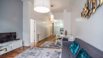 Una sala moderna con un sofá gris, un televisor y una mesita de centro. Hay un pasillo que conduce a una zona de cocina.