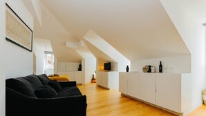 Un espace de vie moderne avec un canapé noir, un plancher en bois, des meubles blancs et un coin cuisine doté de placards blancs et d’un comptoir.