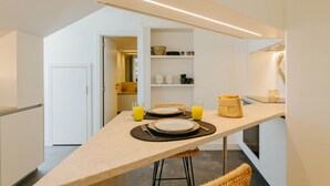Une cuisine moderne dotée d’un grand îlot central, de meubles blancs et d’un four encastré. L’îlot est agrémenté de deux tabourets, et une petite table à manger, avec assiettes et verres, est également présente.
