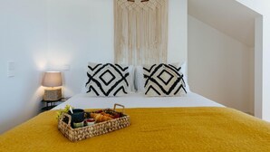 Un lit recouvert d’une couverture jaune, deux coussins à motifs, un plateau en osier contenant de la nourriture et des boissons, une décoration murale en macramé et une lampe sur une table de chevet.