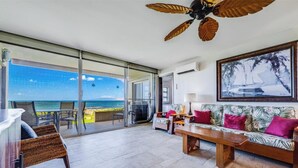 Amplia sala de estar con vista al océano, ventilador de techo, sofá con cojines y una mesa de comedor.