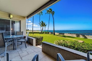 A patio with a table and chairs, overlooking a beach with palm trees and a clear blue sky.