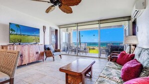 A spacious living area with a large TV, a wooden coffee table, and a sofa with red cushions. There is a ceiling fan and a view of the ocean through large glass doors.