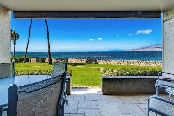 A patio with a view of the ocean, featuring a table and chairs, a stone wall, and a well-maintained lawn.
