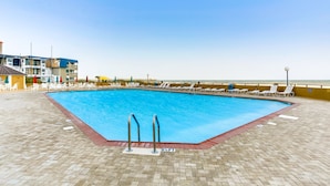 Et rektangulært swimmingpool med to stiger, omgivet af liggestole og parasoller, med en strand og bygninger i baggrunden.