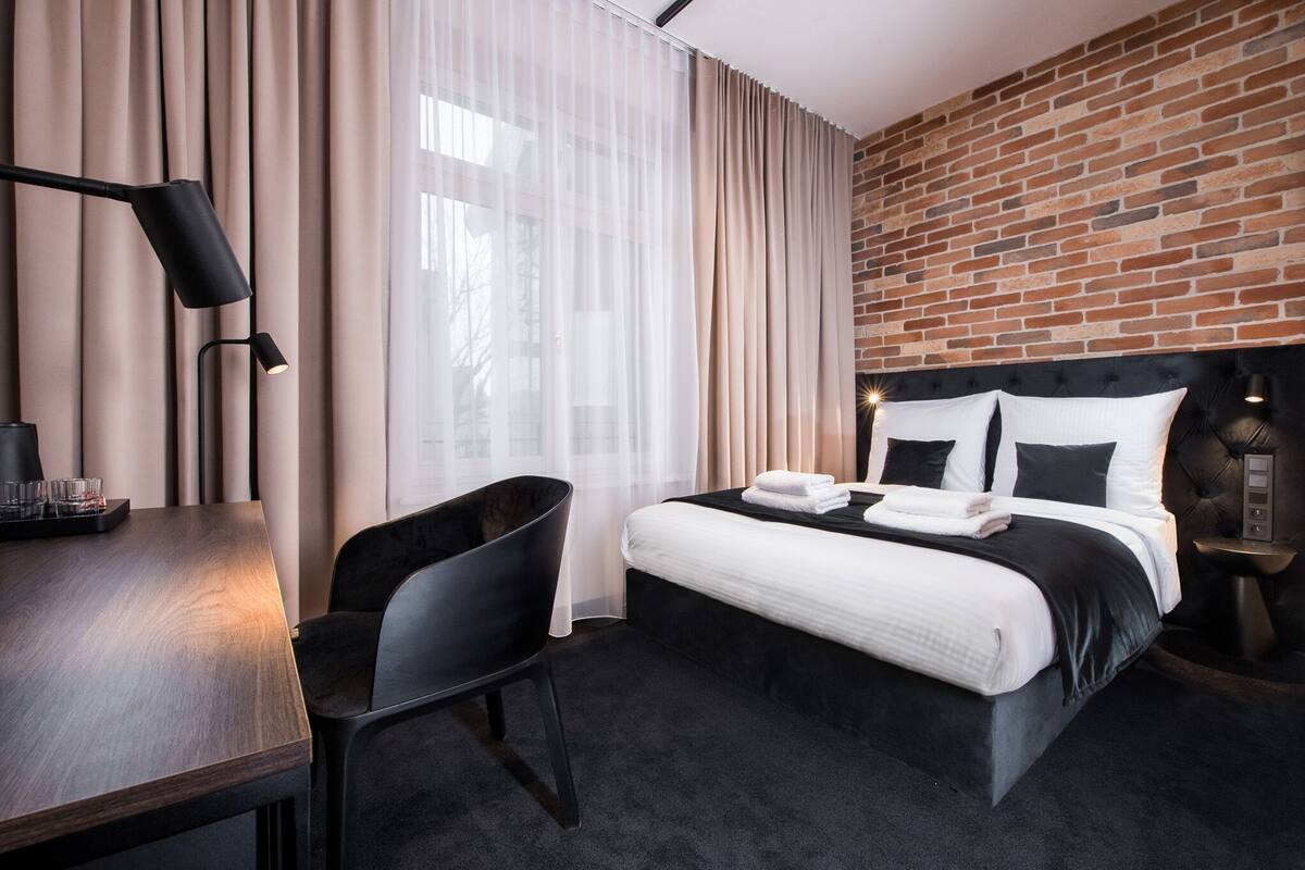 A hotel room with a bed, a desk, a chair, a lamp, and a brick wall.