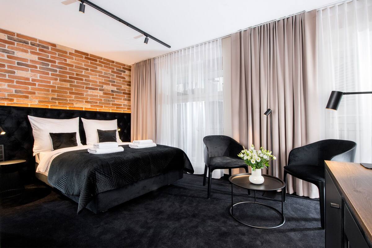 A modern hotel room with a brick accent wall, a large bed with black bedding, two black chairs, a small table with a vase of flowers, and a desk with a lamp.