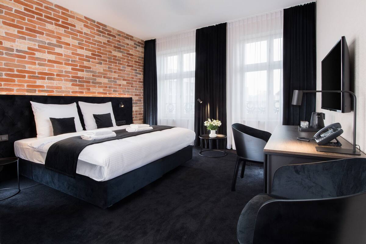 A hotel room with a brick wall, a bed with white bedding, a desk with a chair, a flat-screen TV, and a telephone.