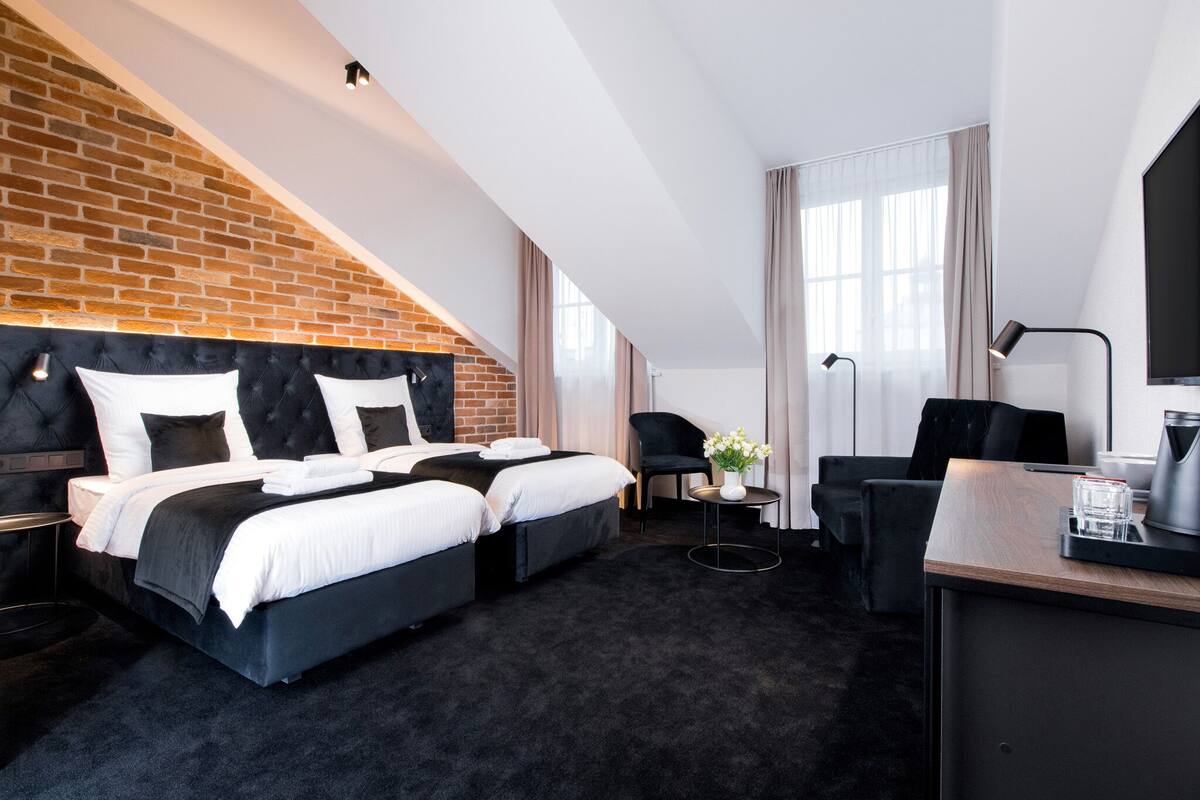 A modern hotel room with a brick accent wall, a large bed with white bedding, two black chairs, a desk with a lamp, and a coffee maker.