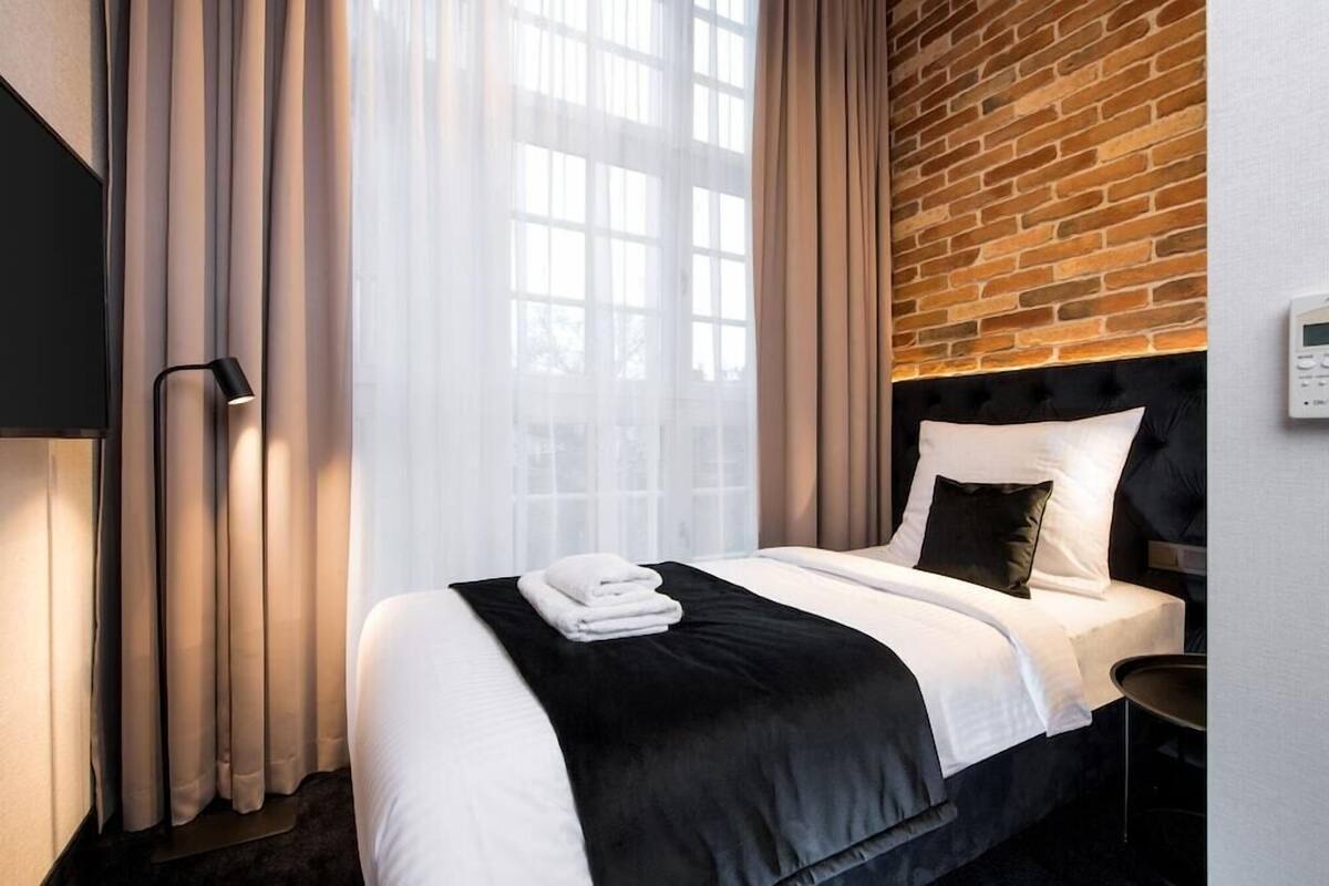 A hotel room with a bed, curtains, a lamp, a thermostat, and a brick wall.