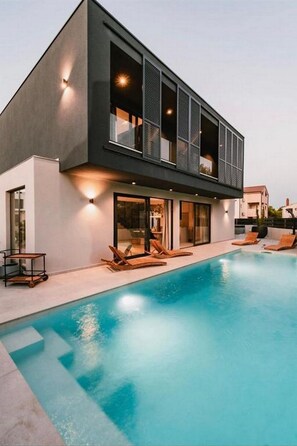Une maison moderne à deux étages, dotée d’une piscine, de grandes fenêtres et d’un espace salon extérieur.