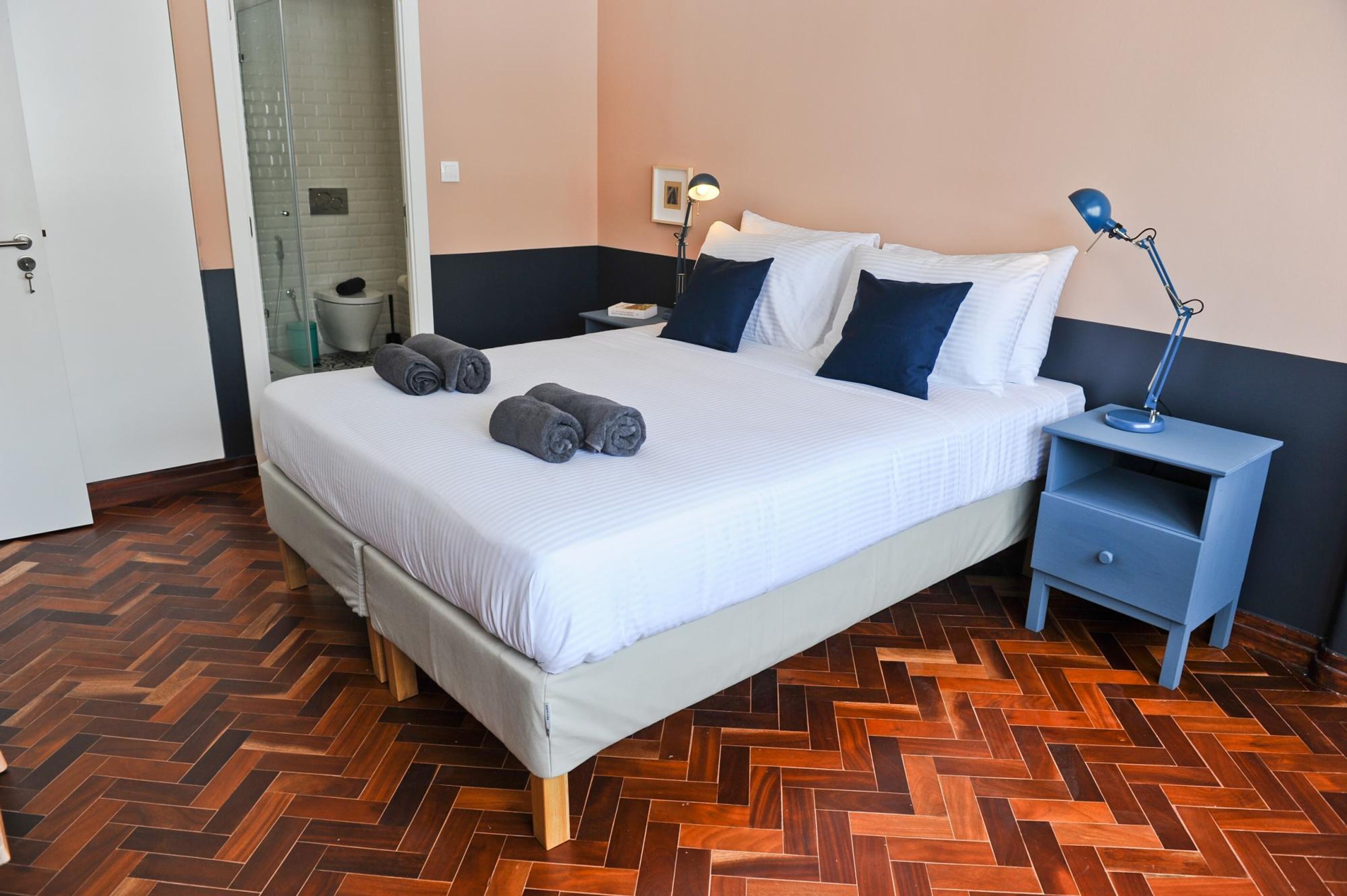 A neatly made bed with rolled towels, a bedside table with a lamp, and a herringbone-patterned wooden floor.