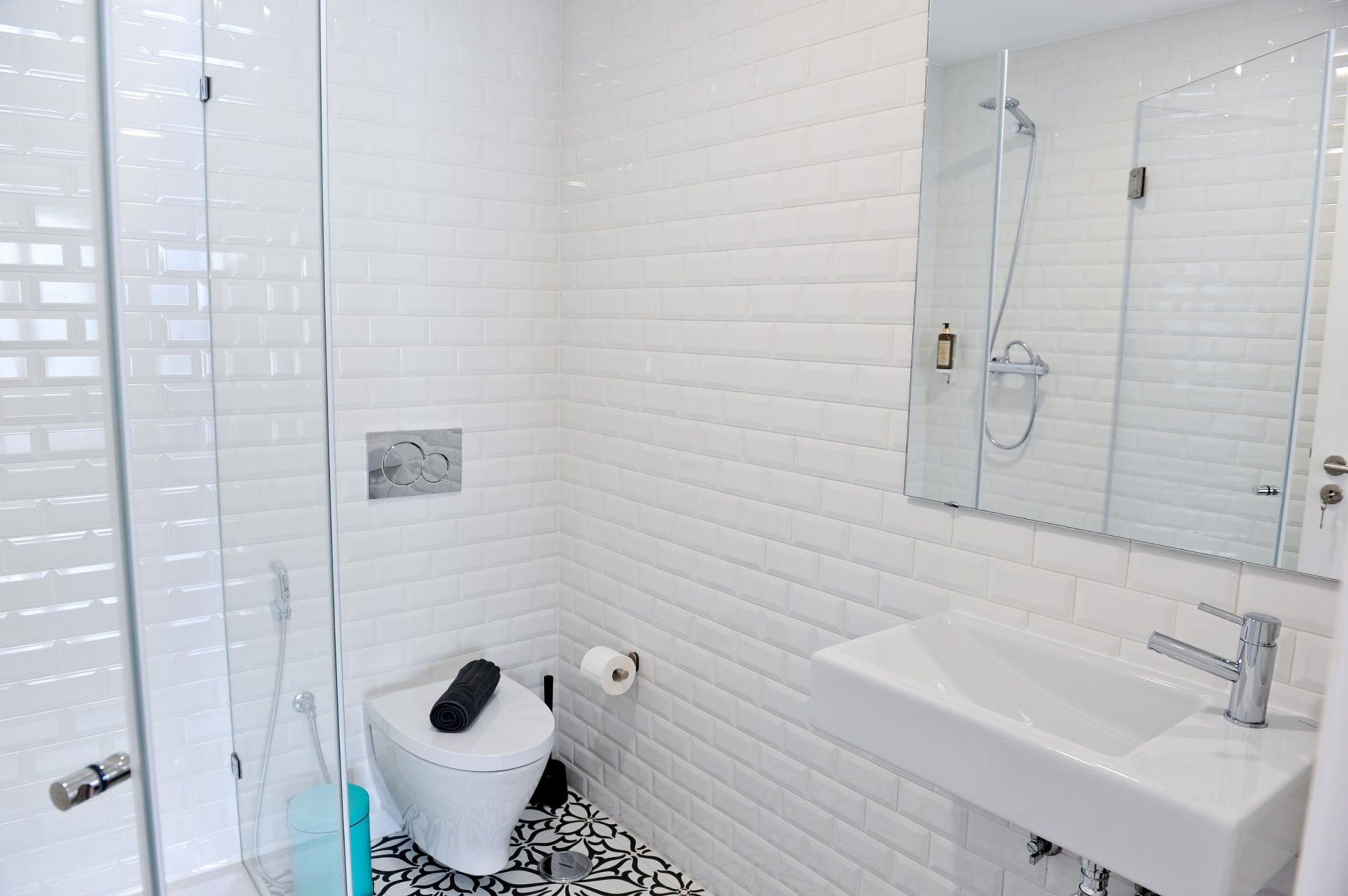 Baño con paredes de azulejos blancos, lavamanos blanco y cabina de ducha de vidrio.