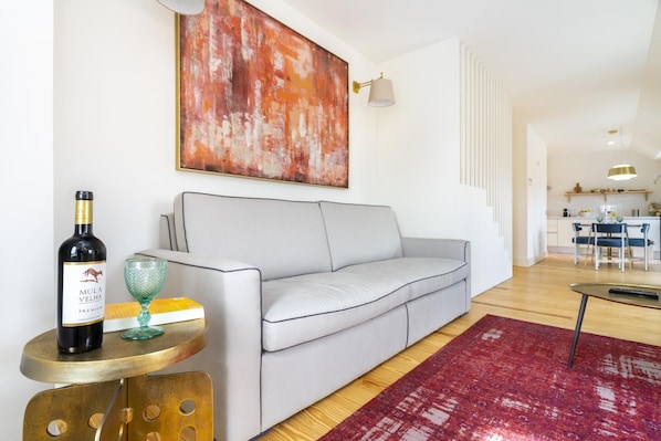 A modern living room with a grey sofa, a red rug, a small round table with a bottle of wine, and a green glass.