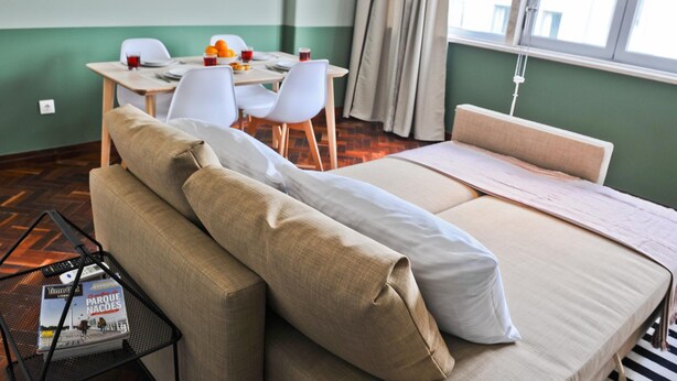 A hotel room with a bed, a small table with a magazine, a chair, and a dining table with chairs.