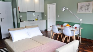A room with a bed, a dining table with chairs, a desk, a lamp, and a kitchen area.