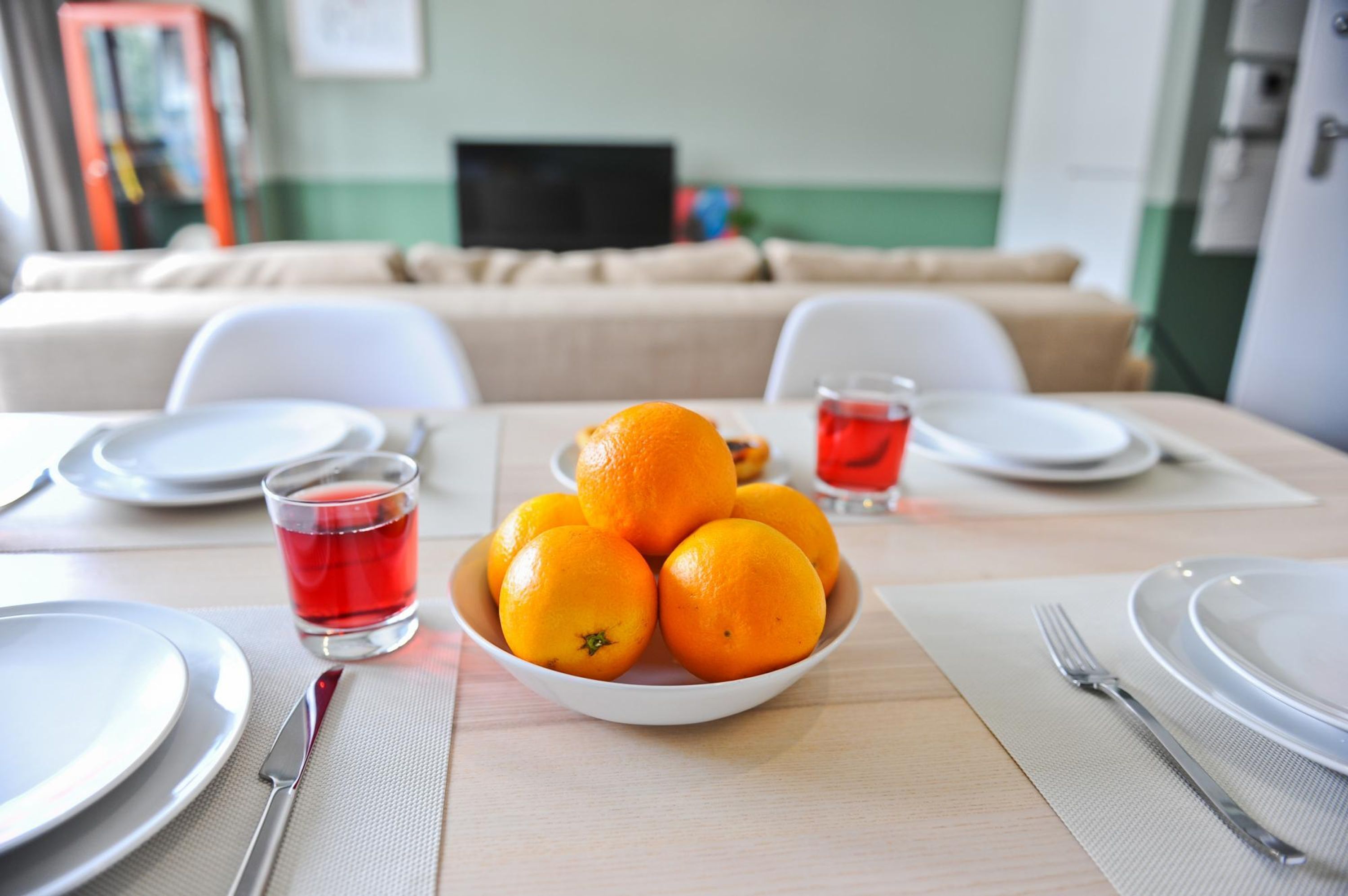 Une table à manger avec un saladier rempli d’oranges, un verre d’une boisson rouge, ainsi que des assiettes et des couverts disposés avec soin.