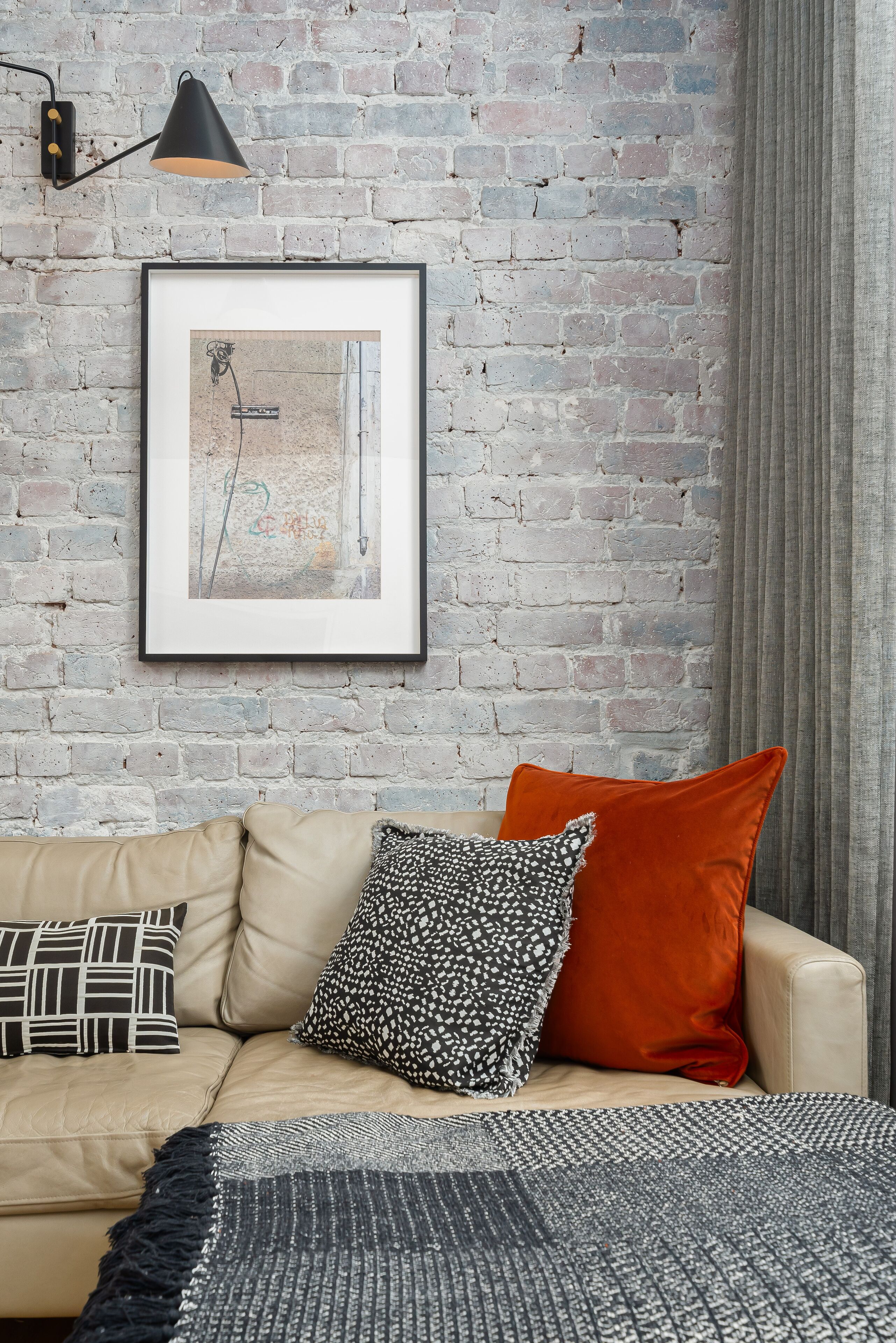 Een kamer met een bakstenen muur, een ingelijst kunstwerk, een beige bank met gekleurde kussens, en een zwarte lamp.