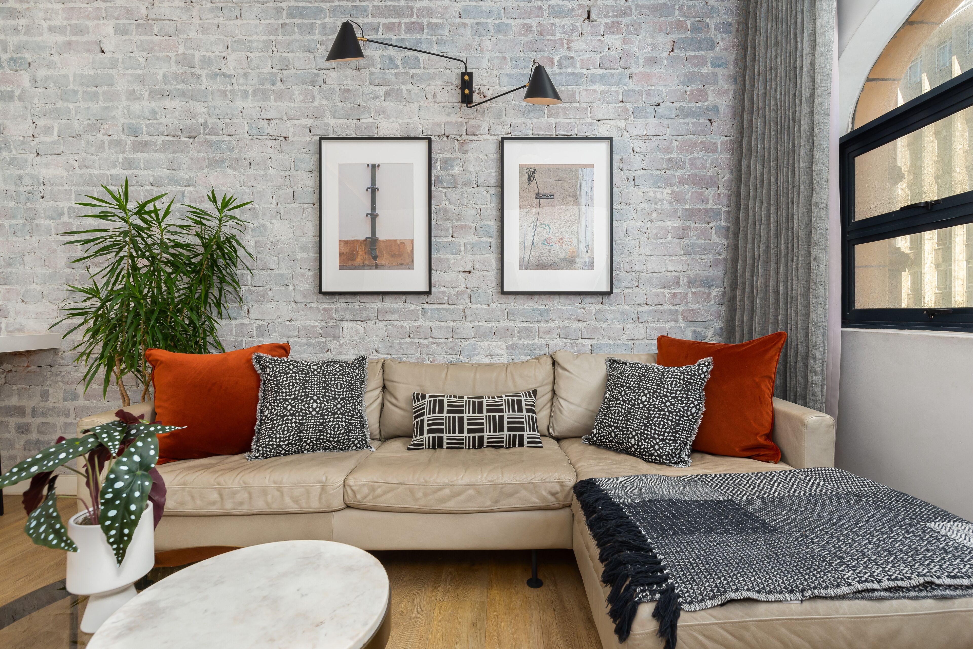 A cozy living room with a brick wall, two framed pictures, a beige sofa with patterned cushions, a round coffee table, and a large plant.