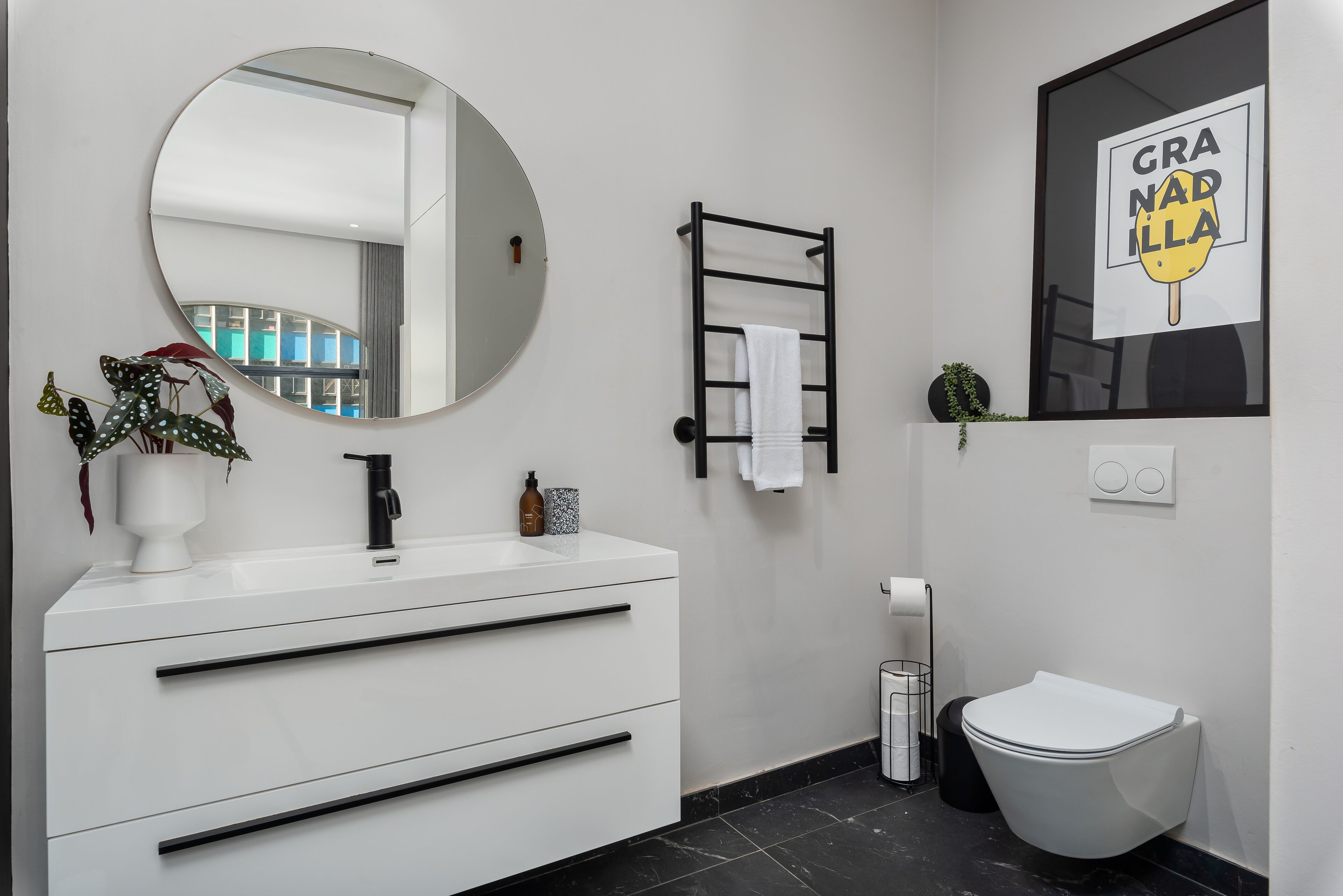 A modern bathroom with a white vanity, a round mirror, a black towel rack, a toilet, and a framed sign.