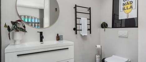 A modern bathroom with a white vanity, a round mirror, a black towel rack, a toilet, and a framed sign.