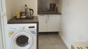 A compact kitchen with a washing machine, a coffee maker, and a kettle on the countertop. - Quiet apartment with balcony (Bois-Colombes)