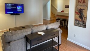 A modern living room with a gray sofa, a dark wooden coffee table, and a flat-screen TV displaying a Roku interface. A staircase leads to a dining area with a wooden table and chairs. A poster on the wall reads "VAIL Colorado Skiing Bliss!"