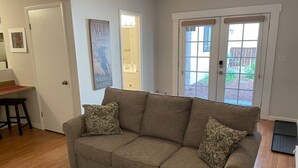 A living room with a beige sofa, two patterned cushions, a wooden coffee table, and a door leading to a backyard.