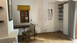 Un dormitorio con cama, una silla, un ventanal con persiana amarilla, una mesita de noche y una estantería con un marco de fotos.