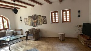 Una sala de estar con una mesa de centro de vidrio, un sofá de mimbre y una chimenea de piedra. Hay dos ventanas con marcos de madera y una obra de arte colgada en la pared.