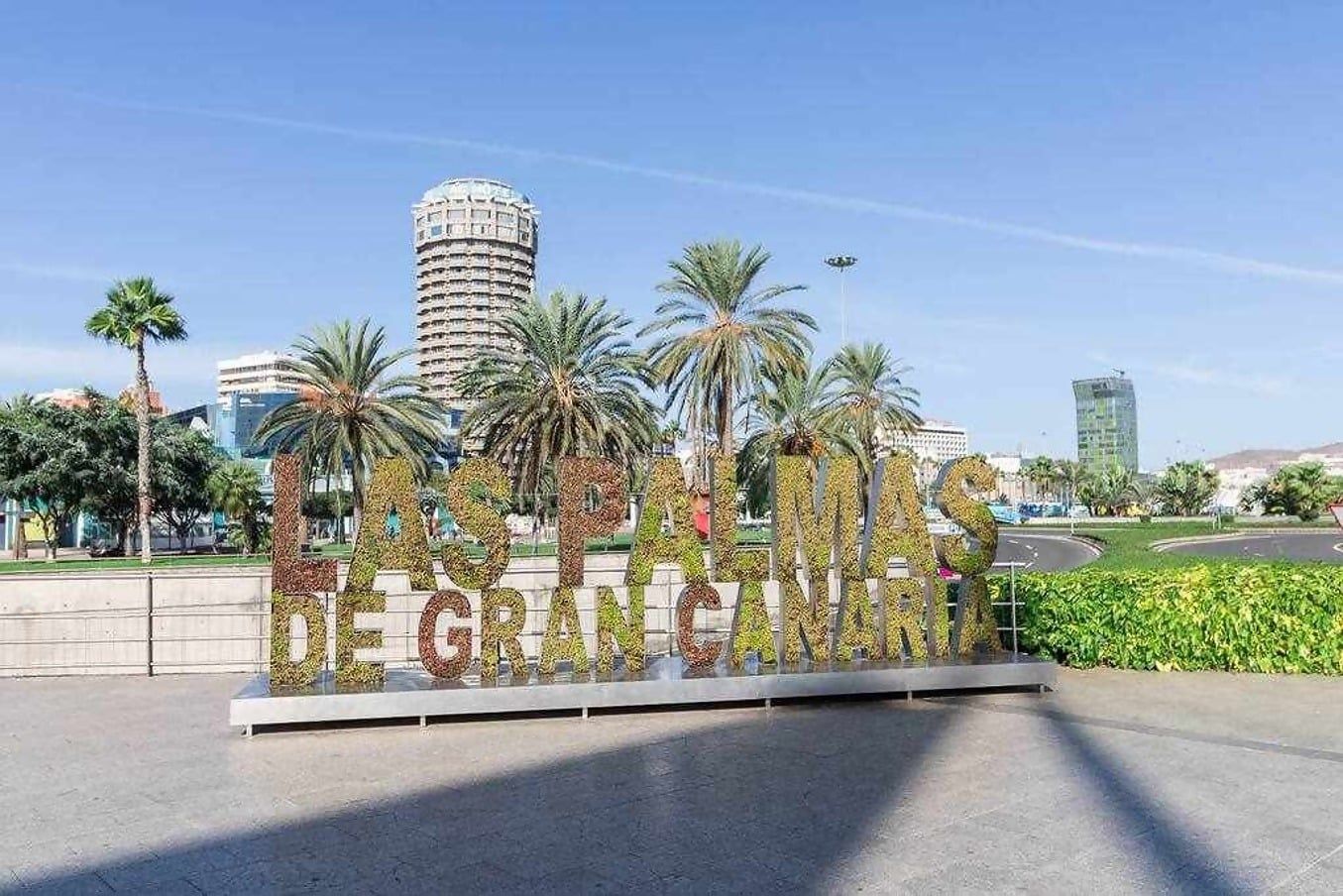 Ein Schild mit dem Text 'Las Palmas de Gran Canaria', umgeben von Palmen und modernen Gebäuden.