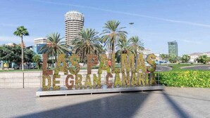 A sign with the text "Las Palmas de Gran Canaria" surrounded by palm trees and modern buildings. - Atamanja Beach Apatment Playa de Las Canteras (Palmas De Gran Canaria, Las)