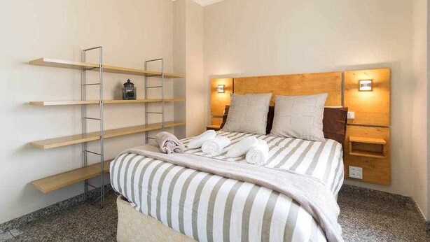 A neatly made bed with striped bedding, wooden headboard, and bedside lamps. There are shelves on the wall and a ladder. - Atamanja Beach Apatment Playa de Las Canteras (Palmas De Gran Canaria, Las)