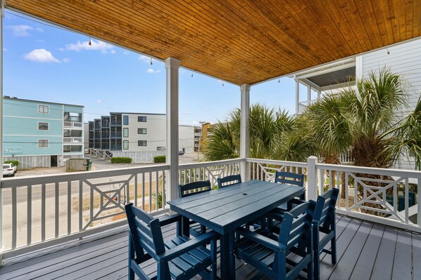 A covered outdoor deck with a table and chairs, overlooking a street with buildings. - 6-Bedroom Home Steps from Garden City Pier with Pool (Garden City Beach)