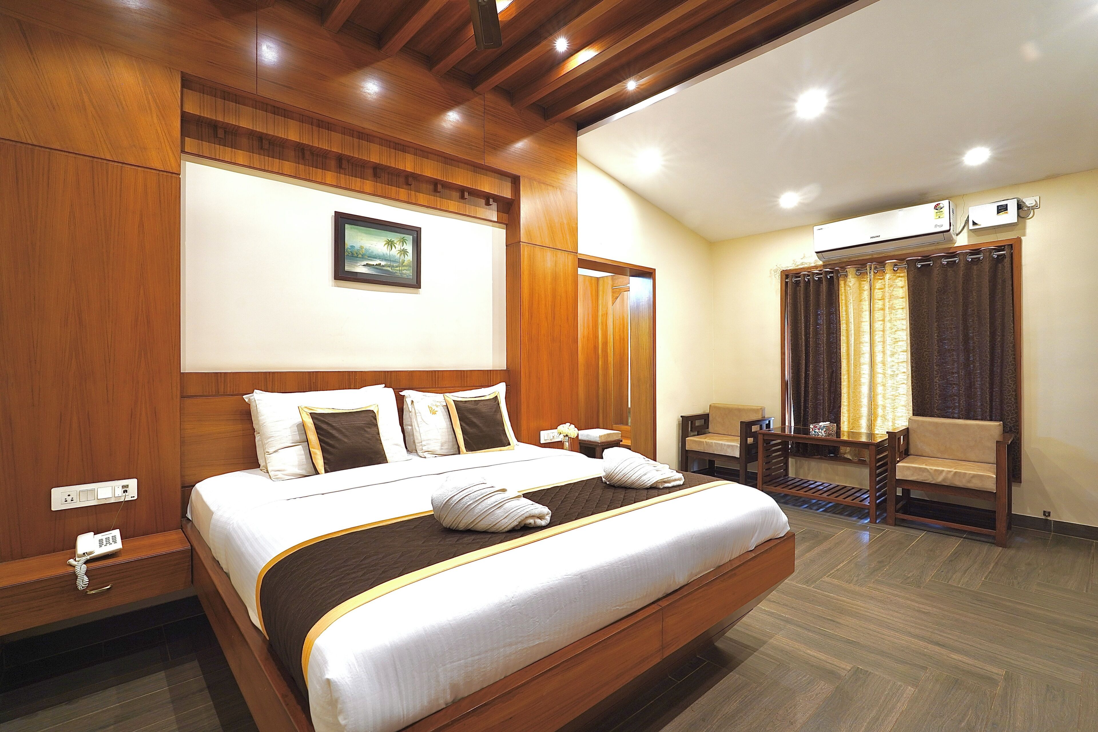 A hotel room with a large bed, wooden paneling, a painting on the wall, a desk with a chair, and an air conditioning unit.